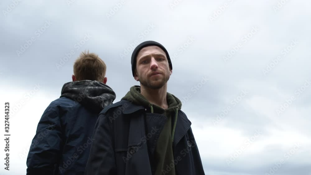 Two Friends Stand With Their Backs To Each Other On The Sky Background. They Look Around. Two Friends Stand With Their Backs To Each Other On The Sky Background. They Look Around.