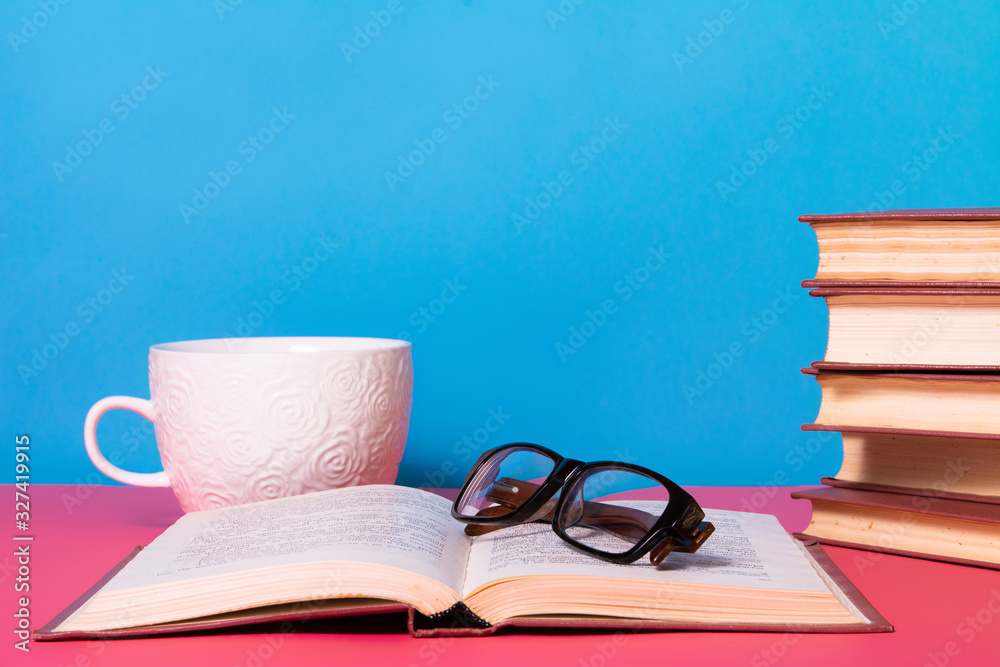 A stack of books with glasses and a mug on a blue and pink background. A stack of books with glasses and a mug on a blue and pink background.