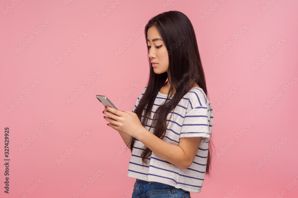 Naklejka premium Side view of girl with brunette hair in t-shirt typing message on cell phone, reading post in social network, chatting online with serious expression. indoor studio shot isolated on pink background