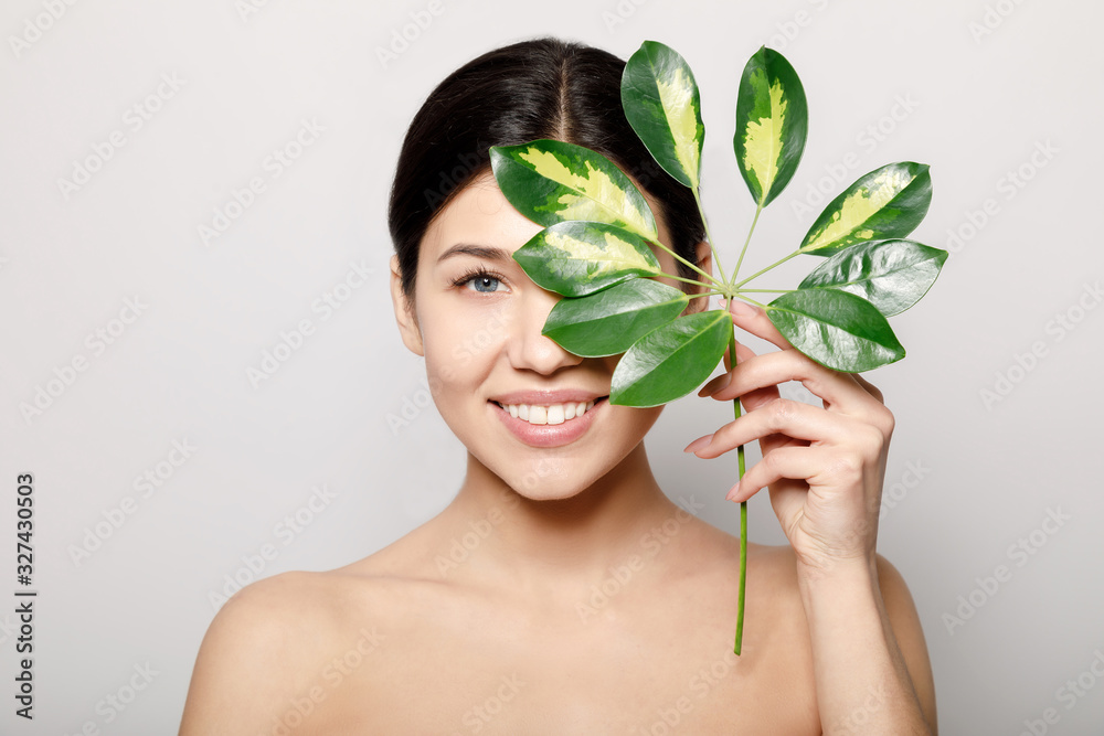 Adorable smiling brunette with perfect skin covering half of face with green leaves isolated light background Adorable smiling brunette with perfect skin covering half of face with green leaves isolated light background