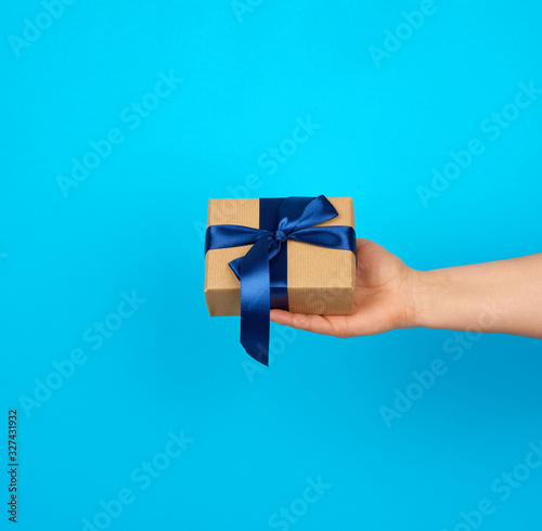 Fototapeta Naklejka Na Ścianę i Meble -  hand holds a box with a gift wrapped in brown kraft paper and tied with a silk blue ribbon