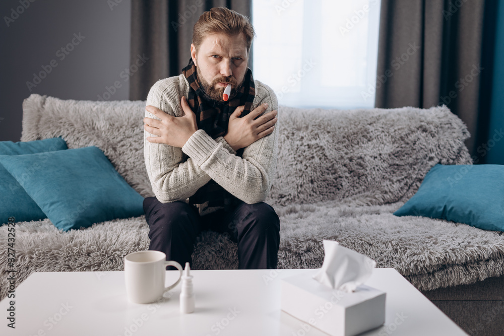 Tired bearded man in warm clothing and knitted scarf measuring body temperature with thermometer while sitting on grey couch. Mature person taking care of himself at home.