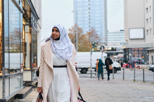 Fototapeta Young stylish Arabic woman in hijab dreamily walking around street with shopping