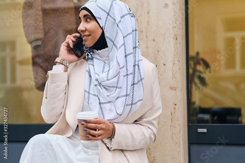 Side view of pretty Arabic woman in hijab with coffee to go talking on smartphone on city street photo