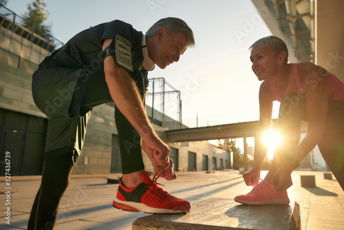 Fototapeta Naklejka Na Ścianę i Meble -  Working out together. Fit and healthy senior couple in sports clothing tying shoelaces before jogging outdoors