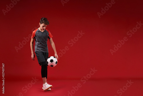 Fototapeta Naklejka Na Ścianę i Meble -  When you play ball, leave it all. A teenage boy engaged in sport, looking at football while training with it. Isolated on red background. Fitness, training, active lifestyle concept. Horizontal shot.