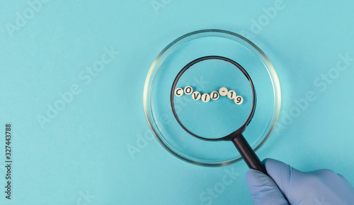 Hand in medical gloves holds a magnifying glass over a petri dish.