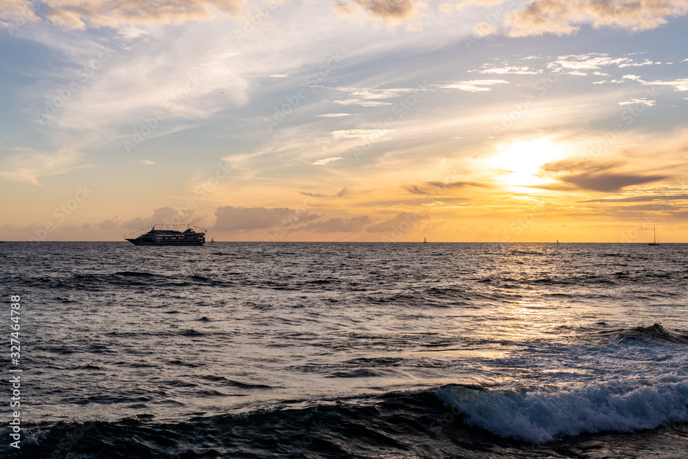 A cruise ship passes in the distance, on the ocean, as sun sets, off an island in the Pacific ocean.