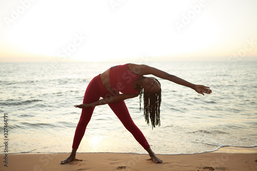 Fit young Black woman standing on sandy beach with sea behind her and stretching right side of body