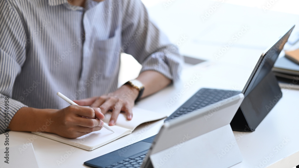 Cropped image of smart man working as Project administrator writing on notebook while sitting in front computer tablet with keyboard case with the modern office as background Cropped image of smart man working as Project administrator writing on notebook while sitting in front computer tablet with keyboard case with the modern office as background