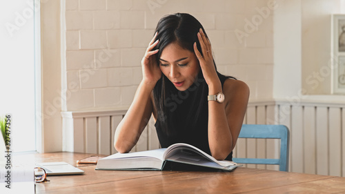 Stressed woman keeping hands on head while reading a book and sitting at the wooden table with comfortable living room as background. © Prathankarnpap