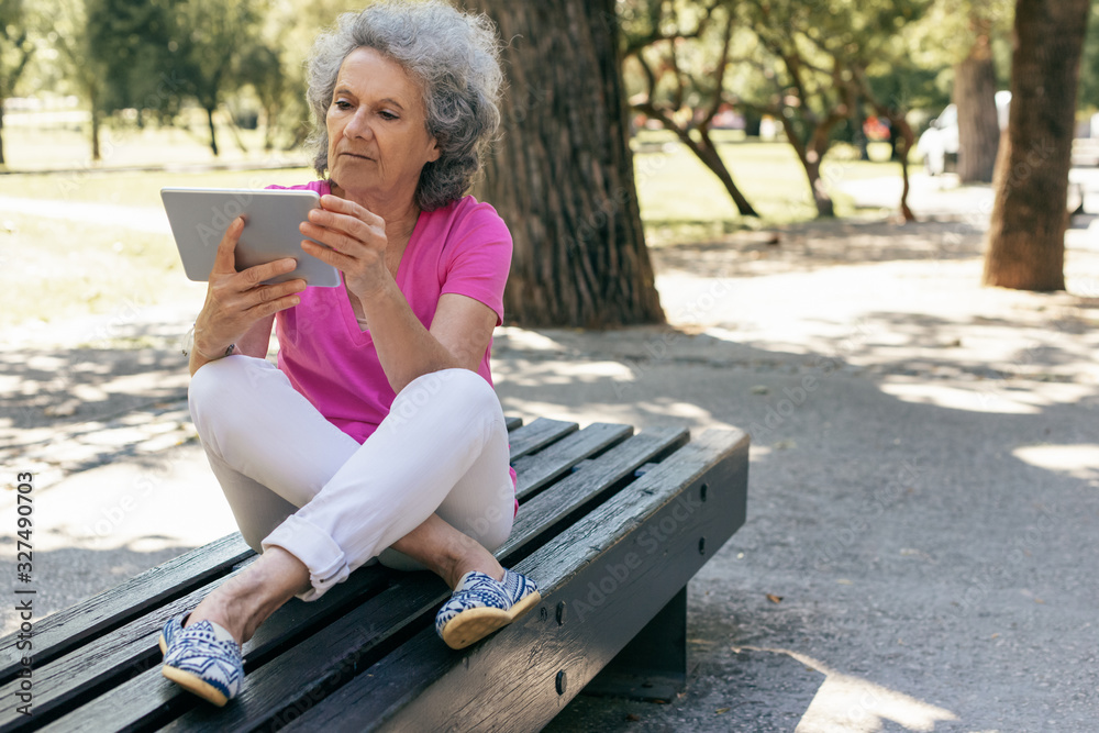 Serious calm old lady watching movie on tablet in park. Senior grey haired woman in casual sitting on park bench and using tablet. Communication outdoors concept