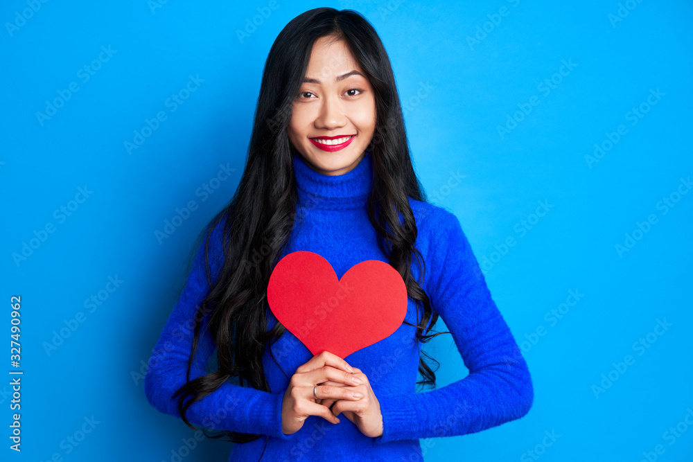 Pretty young woman holding big paper heart in her hands isolated on blue background Pretty young woman holding big paper heart in her hands isolated on blue background