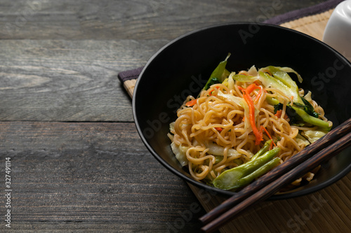 Fried instant noodle with carrot and kale in black bowl on wooden table.