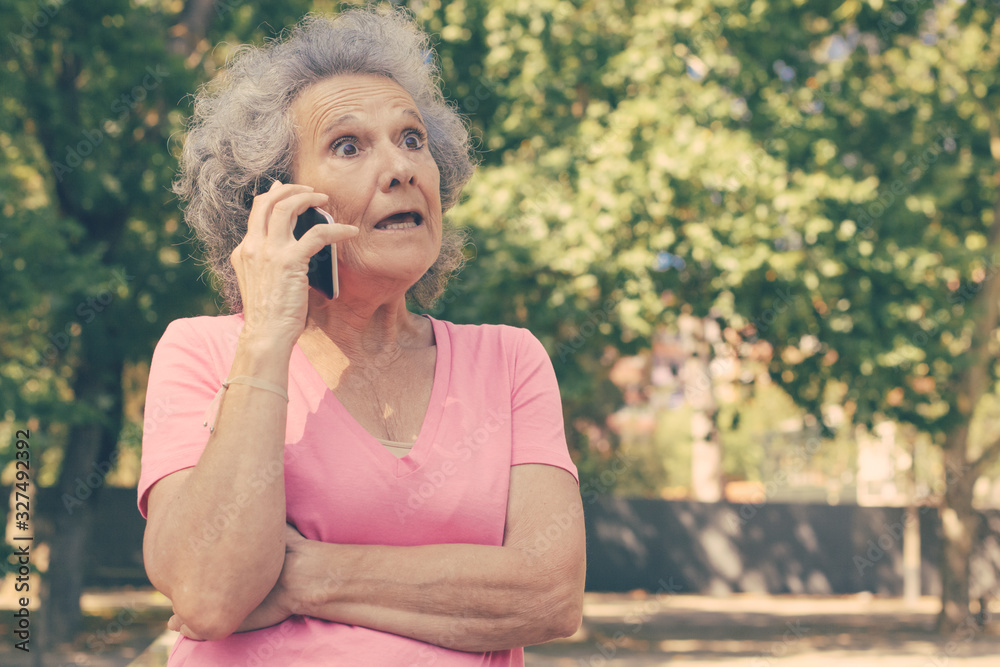 Excited surprised old lady getting shocking news from phone call. Senior grey haired woman with wide eyes standing outdoors and talking on mobile phone. Shock concept Excited surprised old lady getting shocking news from phone call. Senior grey haired woman with wide eyes standing outdoors and talking on mobile phone. Shock concept