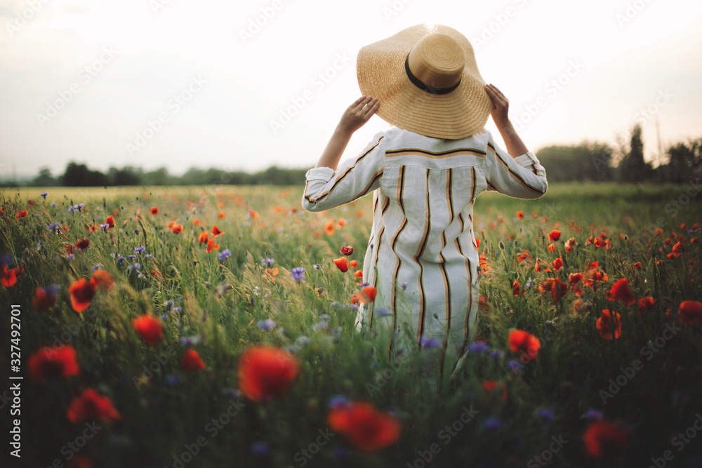 Φωτογραφία Stylish woman in rustic linen dress walking in summer meadow among poppy and wildflowers in sunset light