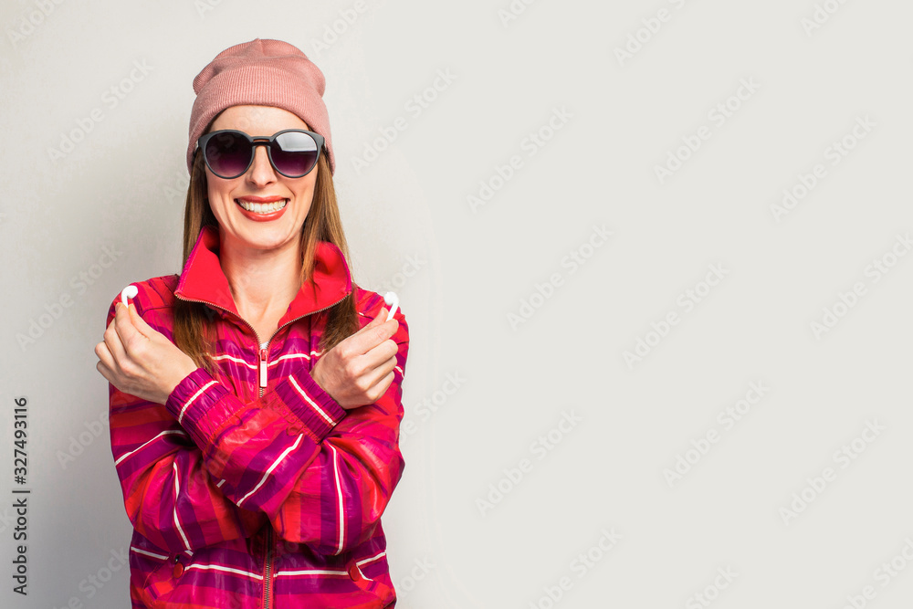 Friendly Young woman in glasses, hat and pink sports jacket with a smiley face holds wireless headphones on a white background. Concept modern style, cool music. Face expression. Banner