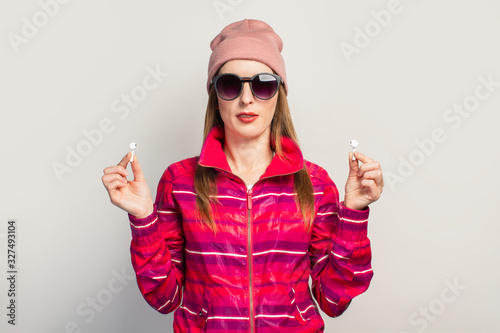 Friendly Young woman in glasses  hat and pink sports jacket with a smiley face holds wireless headphones on a white background. Concept modern style  cool music. Face expression. Banner