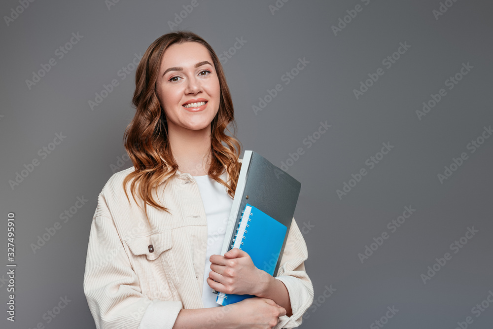 Happy girl student holding a notebook, notepad, a folder in her hands, smiling and looking at the camera on a dark grey background, copy space. Young woman worker, company employee, freelancer Happy girl student holding a notebook, notepad, a folder in her hands, smiling and looking at the camera on a dark grey background, copy space. Young woman worker, company employee, freelancer
