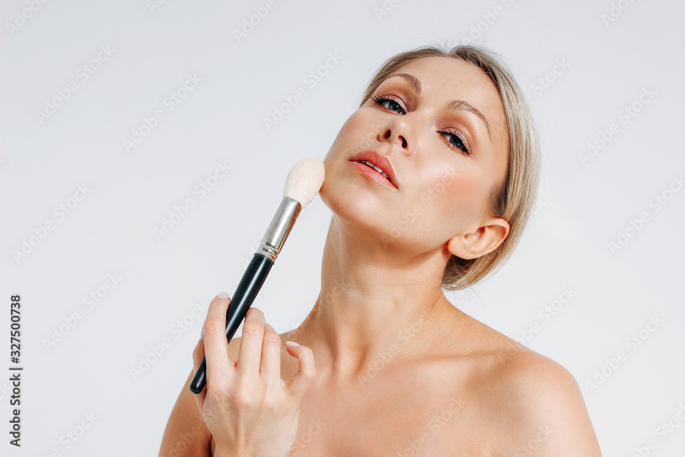 Beauty portrait of blonde smiling woman 35 year plus holding blush brush near clean fresh face isolated on white background