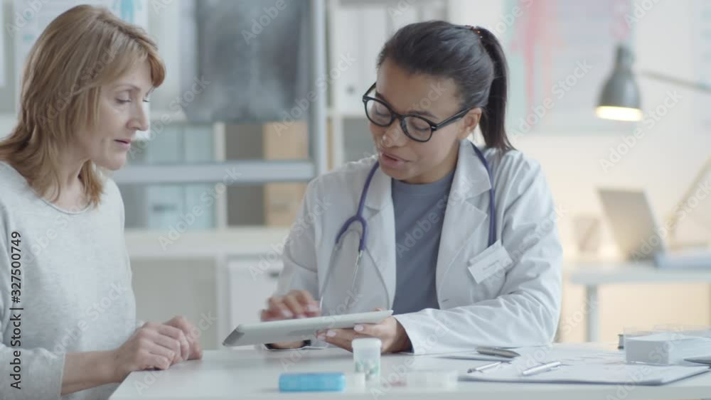 Tracking shot of young cheerful mixed raced female swiping on screen of digital tablet, showing lab test results and talking to middle aged caucasian woman during medical consultation Tracking shot of young cheerful mixed raced female swiping on screen of digital tablet, showing lab test results and talking to middle aged caucasian woman during medical consultation