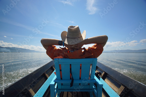 Young man in hat sitting on chair in boat with hands behind head and enjoying life, blue sky and clean water of lake photo