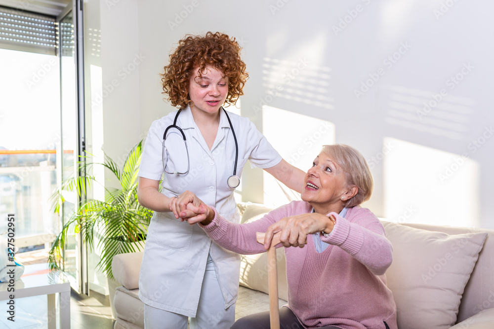 Fototapeta premium Female caregiver helping senior woman get up from couch in living room. Smiling nurse assisting senior woman to get up. Caring nurse supporting patient while getting up from sofa