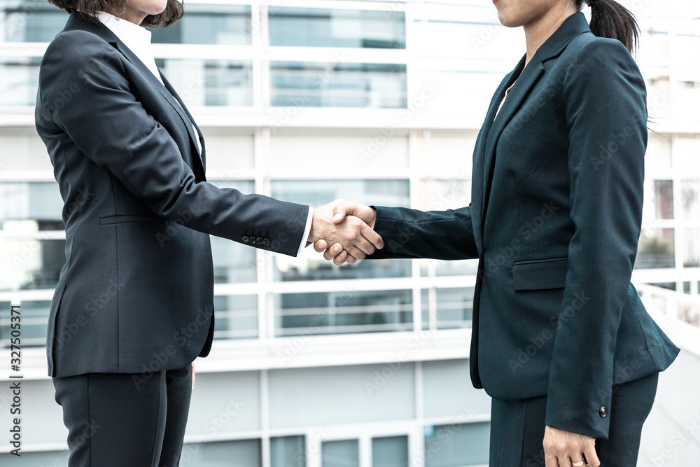 Cropped shot of colleagues shaking hands near office building. Young women wearing formal suits meeting outdoor. Business handshake concept