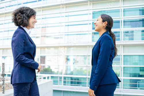 Two smiling colleagues standing on street. Cheerful young confident employees wearing formal suits looking at each other. Business confidence concept