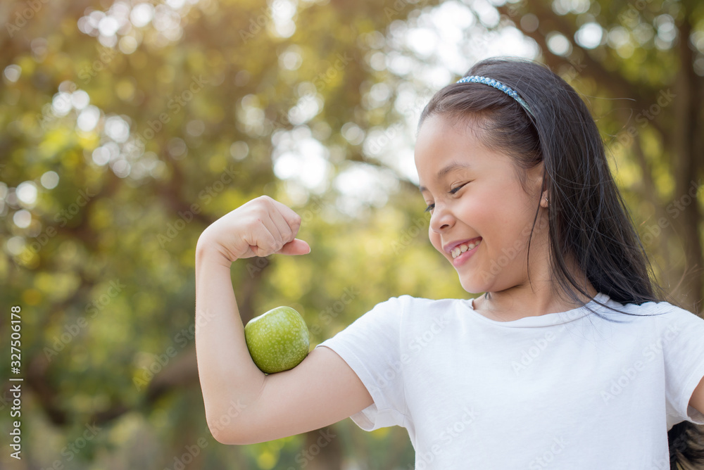 photo happy Little asian girl child standing with big smile. girl with green apple showing biceps..fresh healthy green bio background with abstract blurred foliage and bright summer sunlight photo happy Little asian girl child standing with big smile. girl with green apple showing biceps..fresh healthy green bio background with abstract blurred foliage and bright summer sunlight