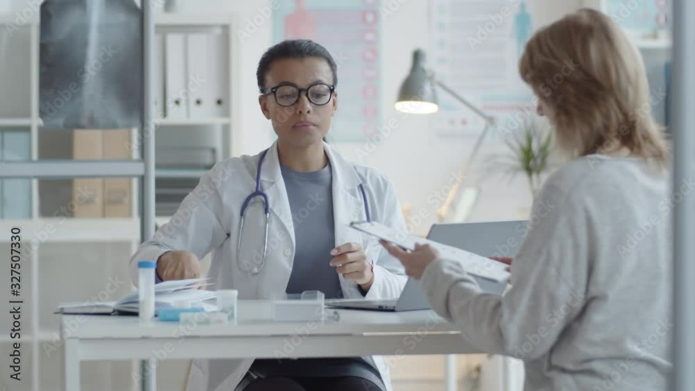 Mixed raced female physician in lab coat and glasses speaking with woman and giving medical service agreement to her for signing while having consultation in clinic Mixed raced female physician in lab coat and glasses speaking with woman and giving medical service agreement to her for signing while having consultation in clinic