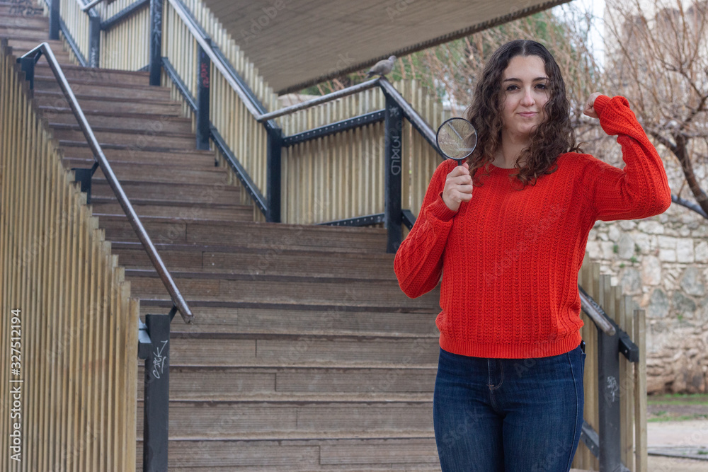 Portrait of rich attractive caucasian young woman with magnifying glass taking a lot of money dollars, in the park, orange sweater and jeans, long curly hair. Place for your text in copy space.