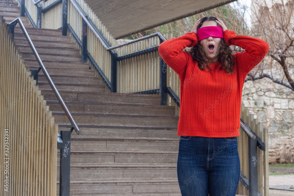 Portrait of attractive caucasian young woman with eye tape being surprised, opening her eyes and mouth with shock, touching her head with hands, in the park, orange sweater and jeans, long curly hair. Portrait of attractive caucasian young woman with eye tape being surprised, opening her eyes and mouth with shock, touching her head with hands, in the park, orange sweater and jeans, long curly hair.