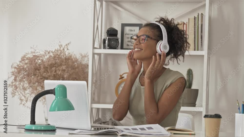Happy pretty african woman in eyeglasses and headphones listening music and dancing while sitting by the table in office