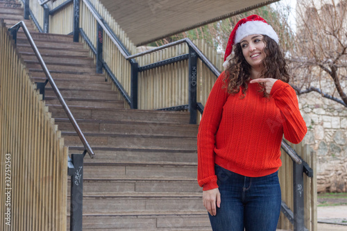 Portrait of attractive young woman model with Santa Claus's hat pointing aside with cheerful expression, showing something amazing at blank space, in the park, orange sweater and jeans, long curly hai