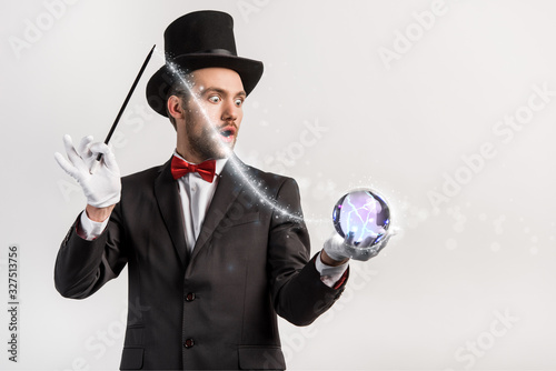 Foto shocked magician holding wand and magic ball isolated on grey with glowing illus