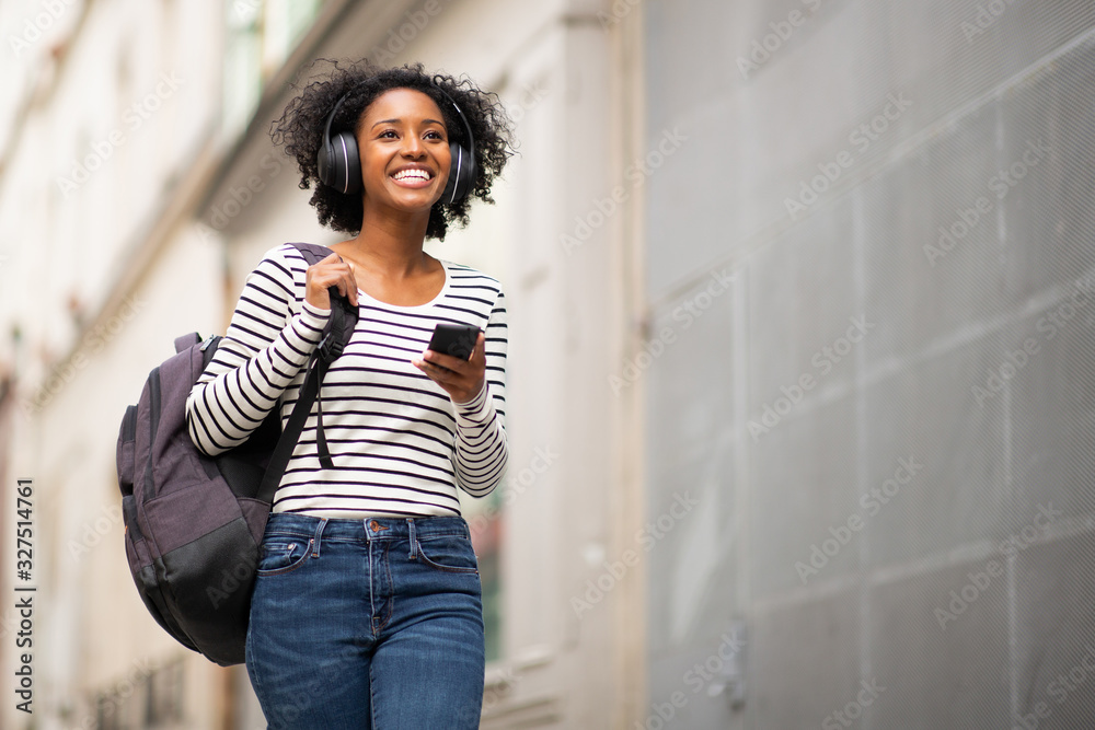 smiling african american woman walking with bag and phone listening to music with headphones in city