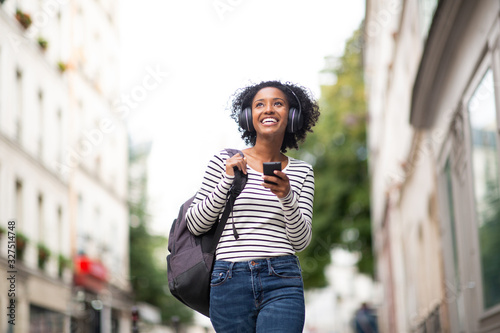 smiling young african american woman walking with mobile phone listening to music with headphones in city