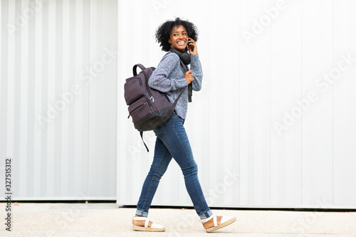 Full length portrait young african american woman walking and talking with mobile phone by white wall
