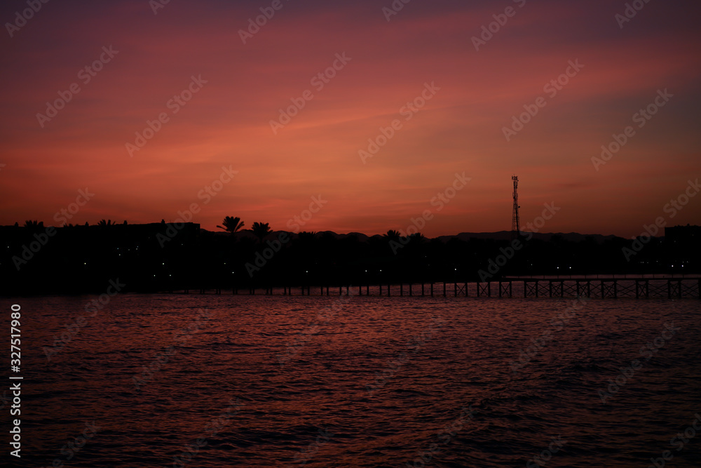 Purple sunset against the background of the silhouette of a pontoon bridge, hotel and palm trees at dusk.