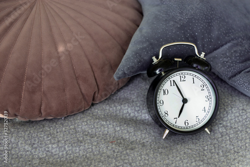 Fotografie Black alarm clock in a made-up bed on a gray textured bedspread, against a pink and blue velor pillow