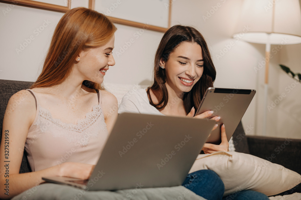 Ginger smiling girl is looking at her brunette friend tablet while sitting on the sofa and having joy together