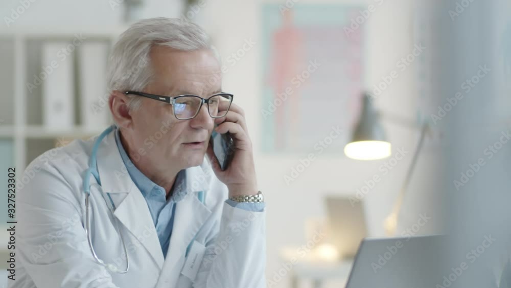 Tilt down shot of senior male doctor browsing the Internet on laptop and having mobile phone conversation while working at desk in clinic