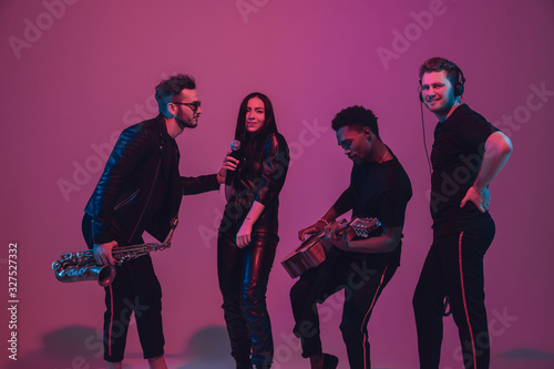 Group of young multiethnic musicians created band, dancing in neon light on pink background. Concept of music, hobby, festival, wellness. Joyful party host, dancer, singer, guitarist, saxophonist. © master1305