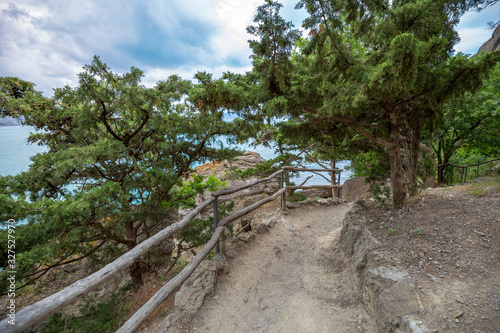 Mountain path fenced by a wooden fence with a view of the sea through the branches of mountain pines and junipers. The Golitsyn trail, Crimea. Rocky coast of the black sea. Seascape.