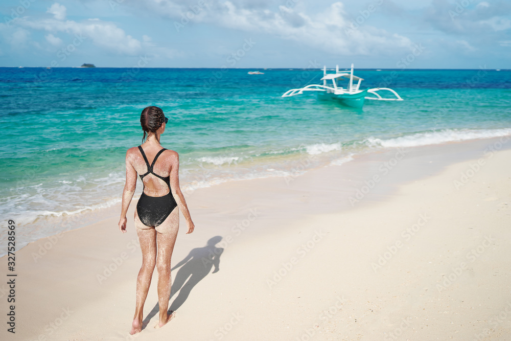 Vacation on the seashore. Back view of young woman walking away on the beautiful tropical white sand beach.