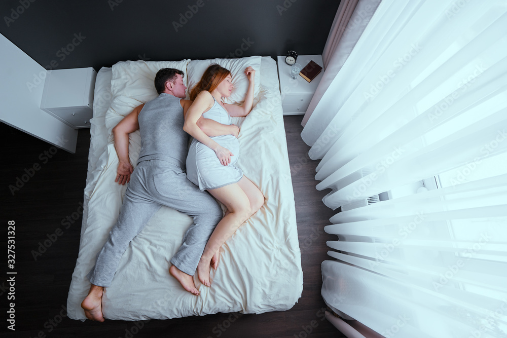 Family couple sleeping together in bed. Husband hugging his pregnant wife. - obrazy, fototapety, plakaty 
