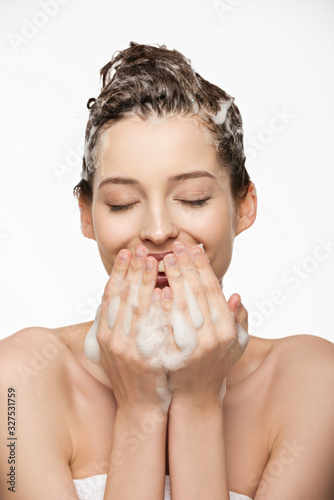 cheerful girl with closed eyes covering mouth with hand while washing hair isolated on white