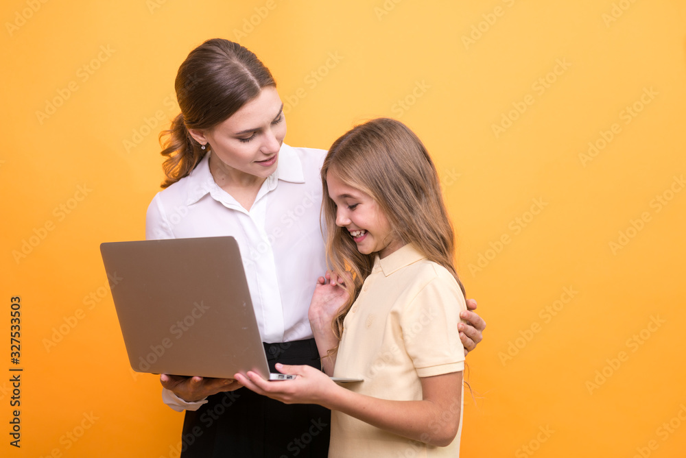 © valerii kalantai - Beautiful teacher and cute schoolgirl hold in hands laptop for study on orange background.
