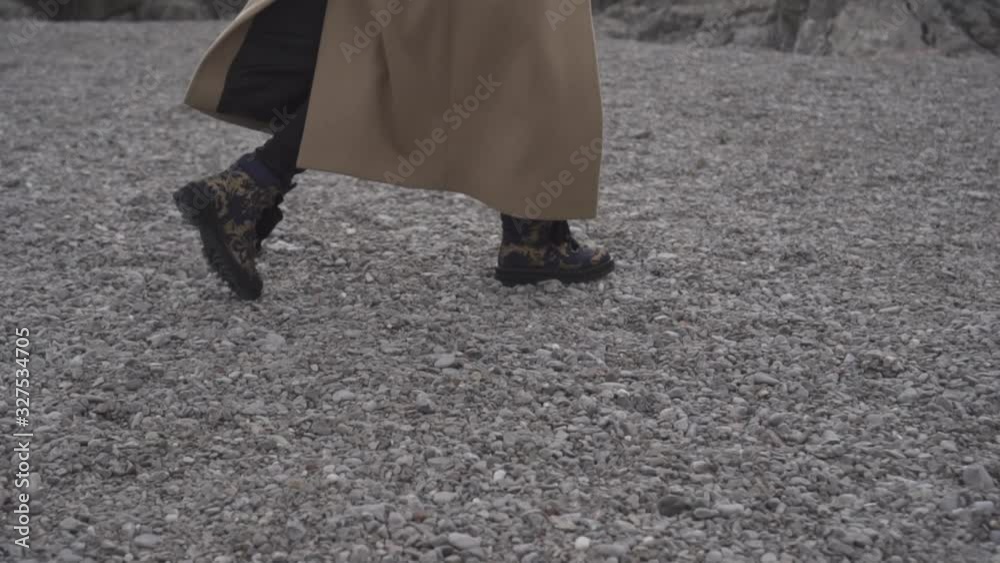 A girl in a beige coat, black jeans and leather boots walks on a rocky seashore A girl in a beige coat, black jeans and leather boots walks on a rocky seashore